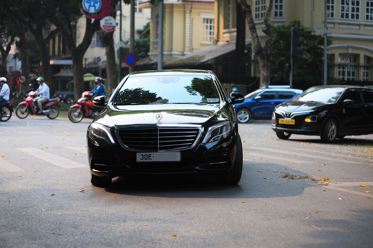 A sleek black car navigates a lively urban street with motorbikes and architecture.